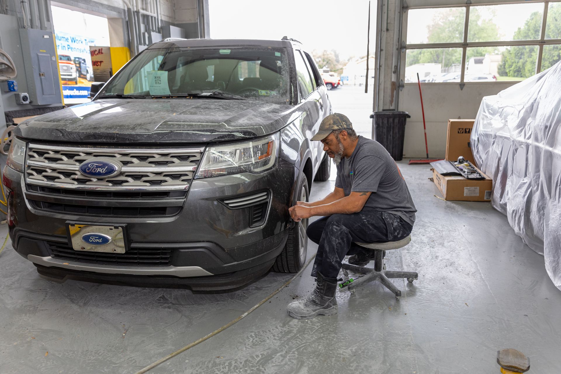A mechanic works on a dark SUV in a garage, seated on a rolling stool.