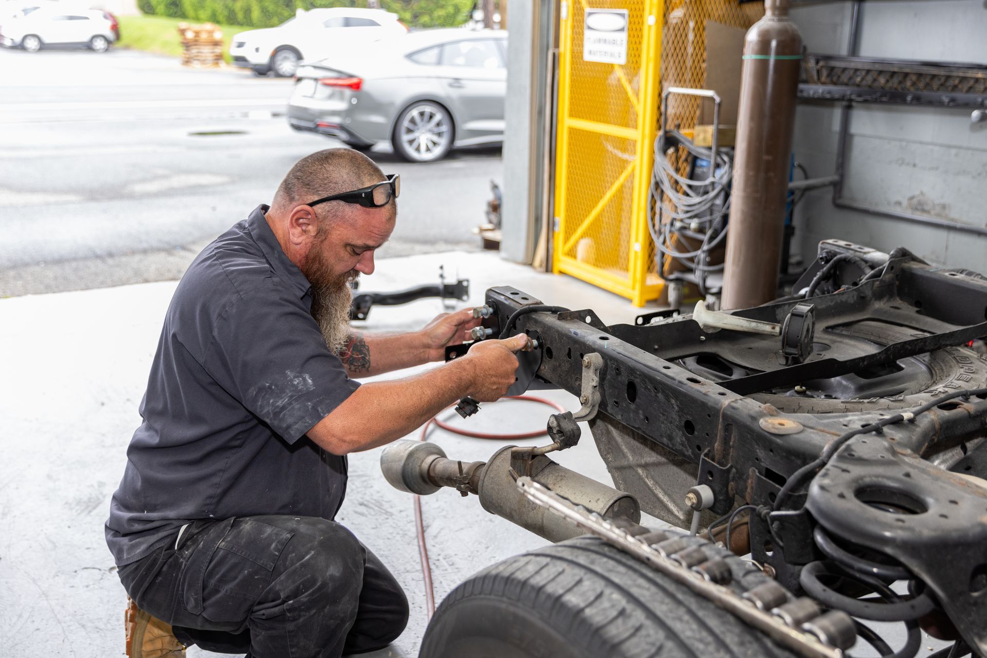Mechanic works on a vehicle chassis outdoors. He wears safety glasses, a black shirt, and kneels while working.