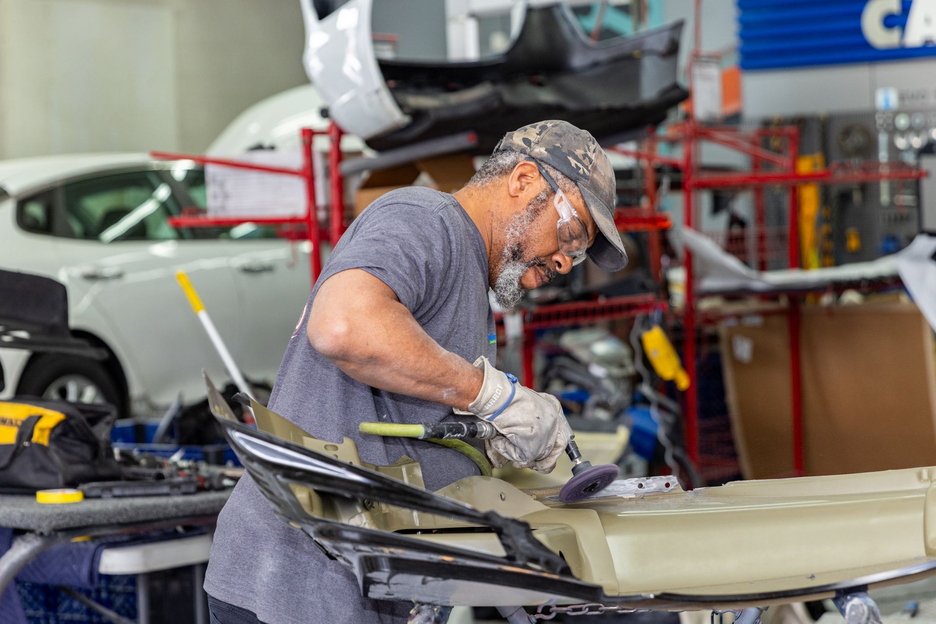 Auto body worker sanding a car bumper in a repair shop; wearing safety glasses and gloves.