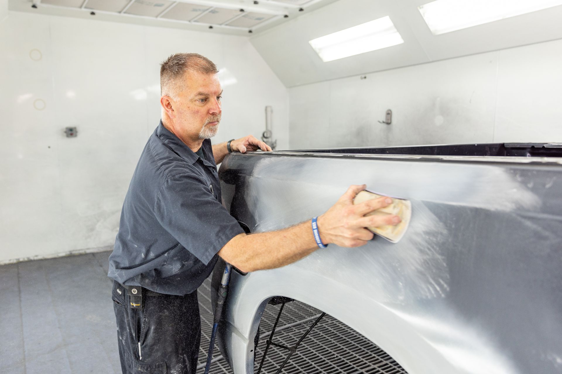 Man sanding a gray truck in a bright auto body shop.
