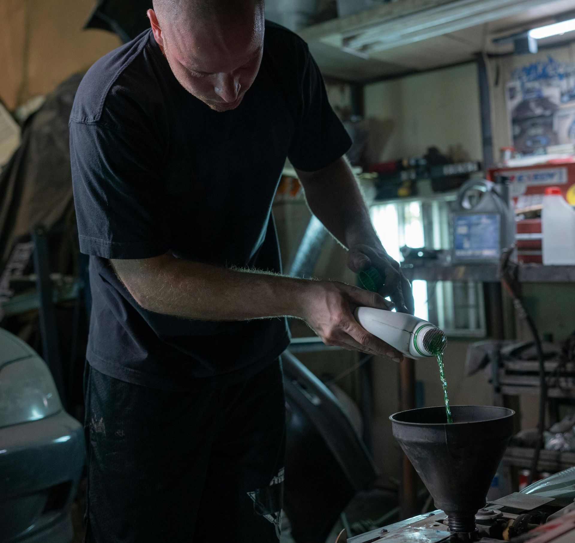 A person wearing a dark t-shirt pours bright green liquid from a bottle into a funnel inside a workshop.