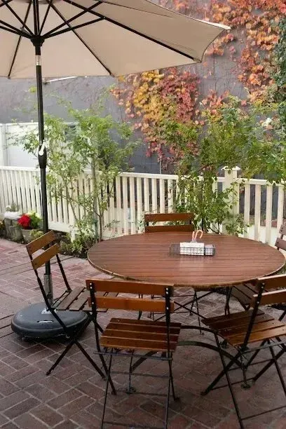 tree service close to me-Outdoor cafe table with chairs under an umbrella, surrounded by a brick patio, white fence, and fall foliage.