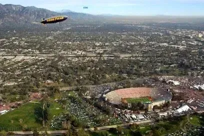 Rose Bowl stadium with a blimp overhead, viewed from above, mountains in background, crowd gathered.