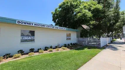 Tree service close to me-Downey History Center Museum: white building with green trim, sign, two windows, grass and sidewalk.