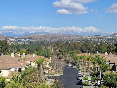 Residential street with houses, trees, and snowy mountains in the background under a blue sky.
