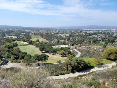 Overlook of a green golf course, trees, and buildings, with a dirt path in the foreground.