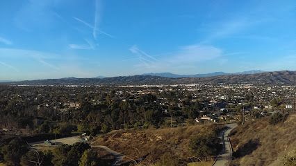Scenic overlook of a city, trees, and mountains under a blue sky with contrails.