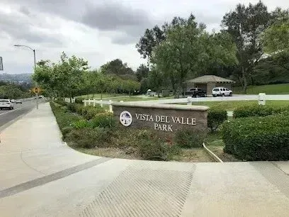 tree service close to me-Vista del Valle Park sign by a road with trees and a cloudy sky.