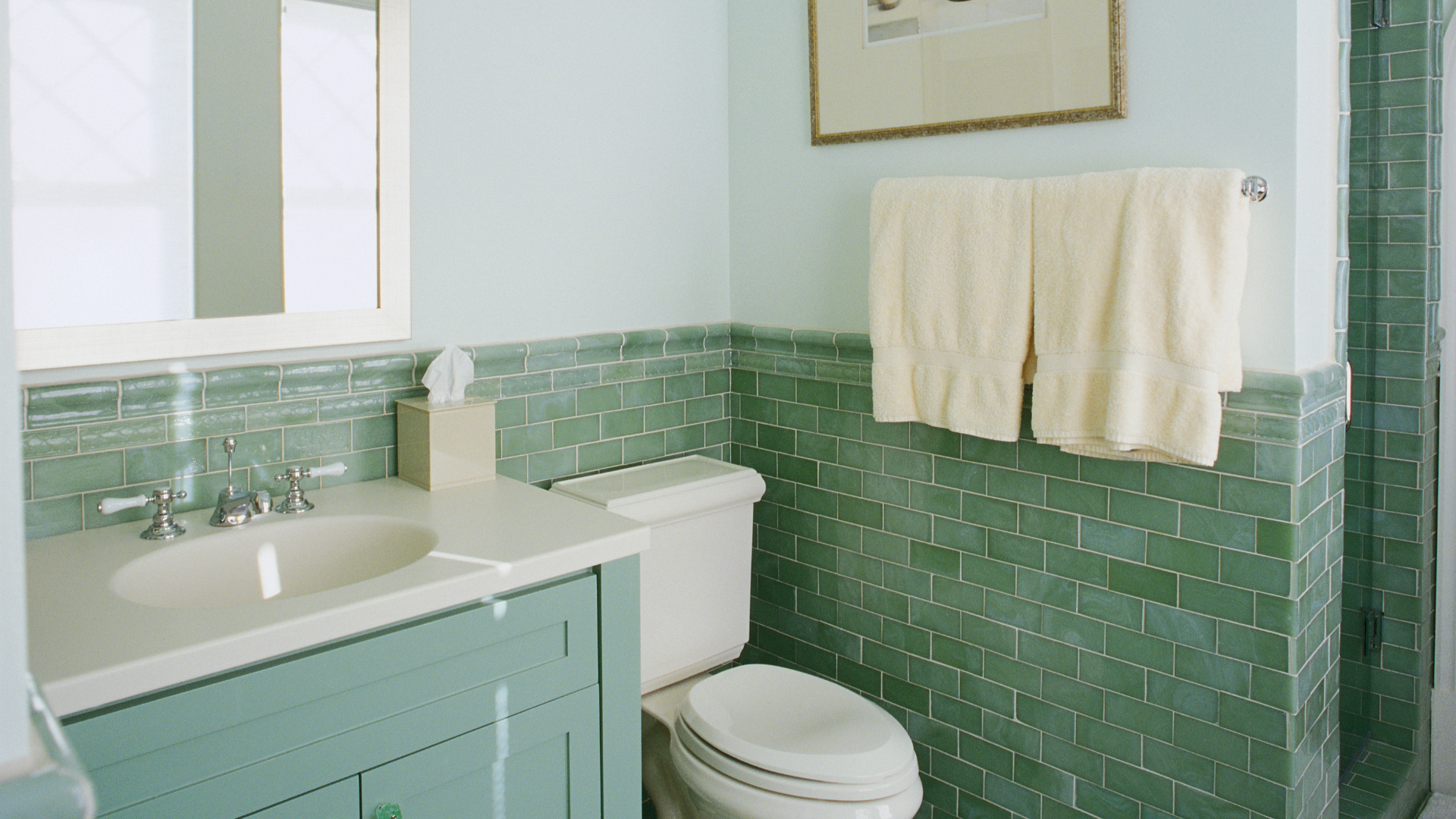 A bathroom with green tiles , a toilet , sink and mirror.