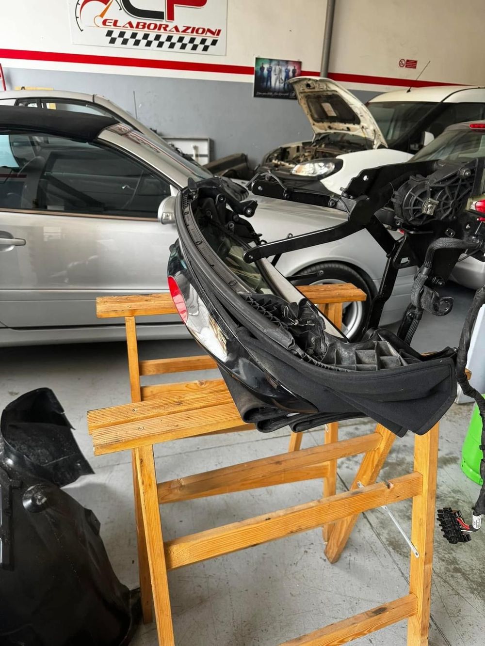 Paraurti anteriore di un'auto smontato su un supporto di legno all'interno di un'officina. Altri veicoli sullo sfondo.