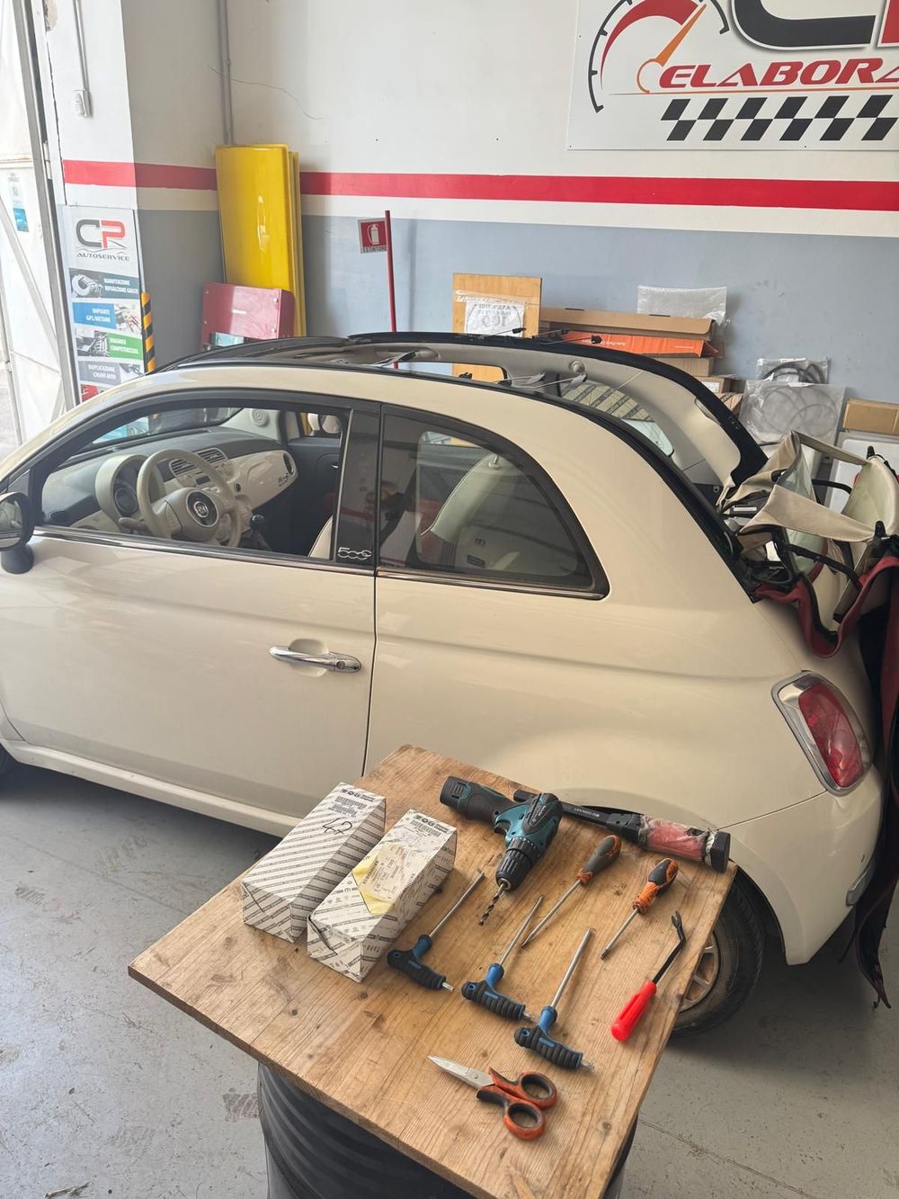 Fiat 500 bianca decappottabile in garage con attrezzi su una superficie in legno. Tettuccio apribile aperto, attrezzi sistemati.