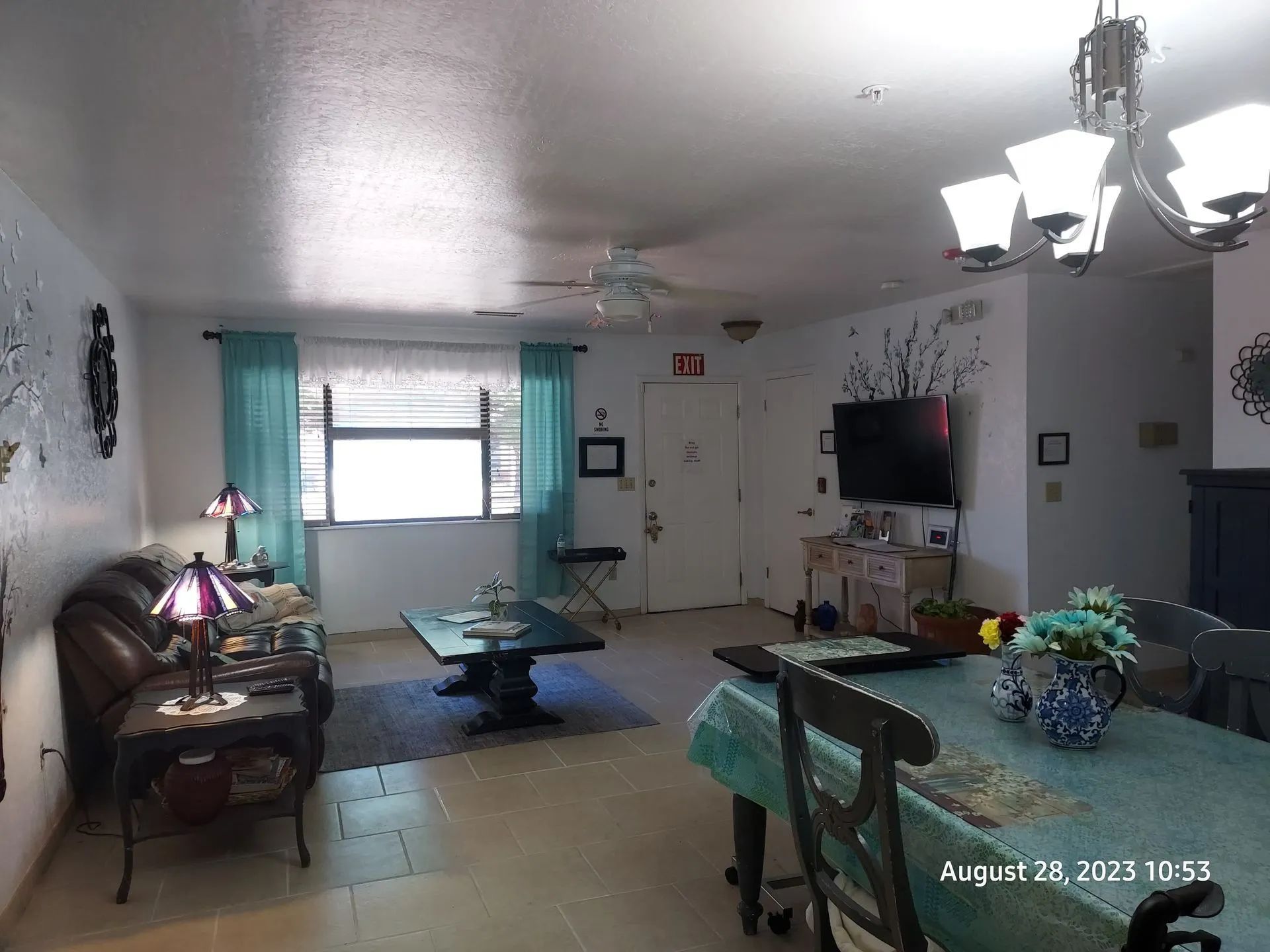 Living room with teal curtains, dark furniture, and floral accents. A TV is mounted on the wall above a small console table.