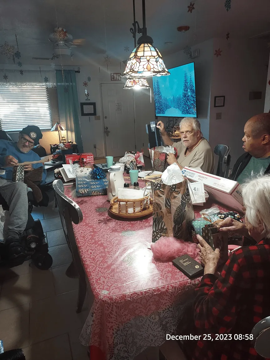 Family at a Christmas table with gifts and food, a few people are holding cameras or taking pictures, the room is decorated.