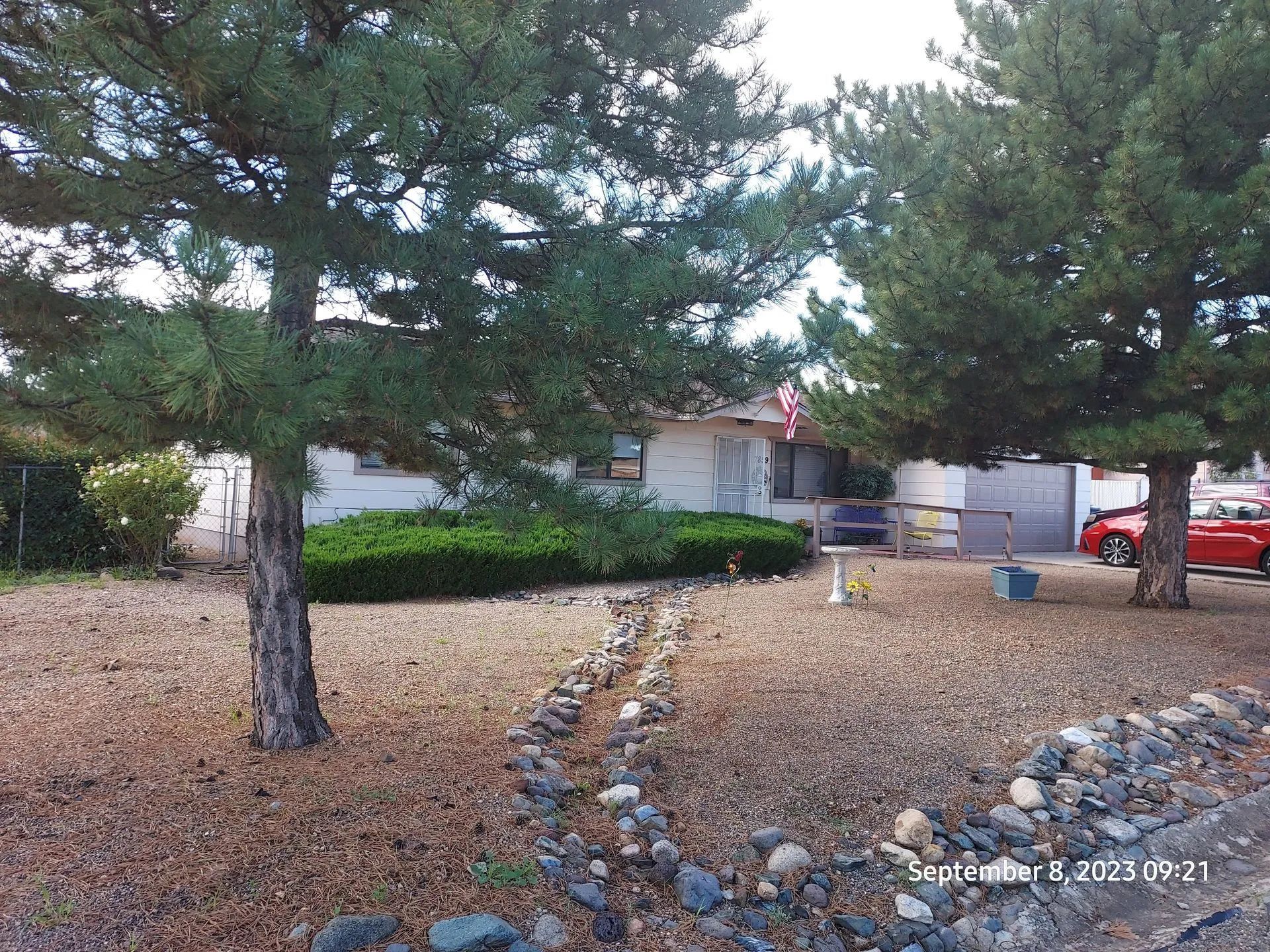 A house with brown landscaping in front, framed by pine trees. A red car sits in the driveway on the right.