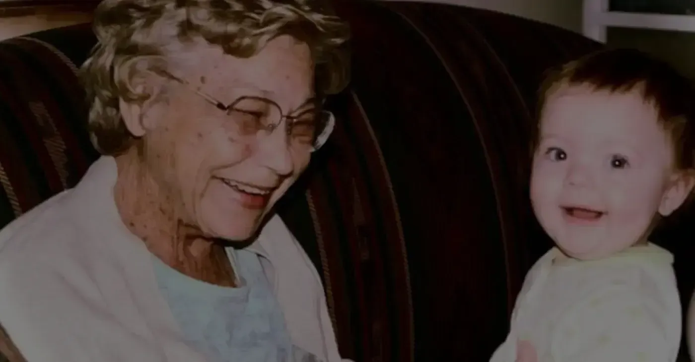 Elderly woman with glasses smiles at a young child with Down syndrome, both sitting on a brown couch.