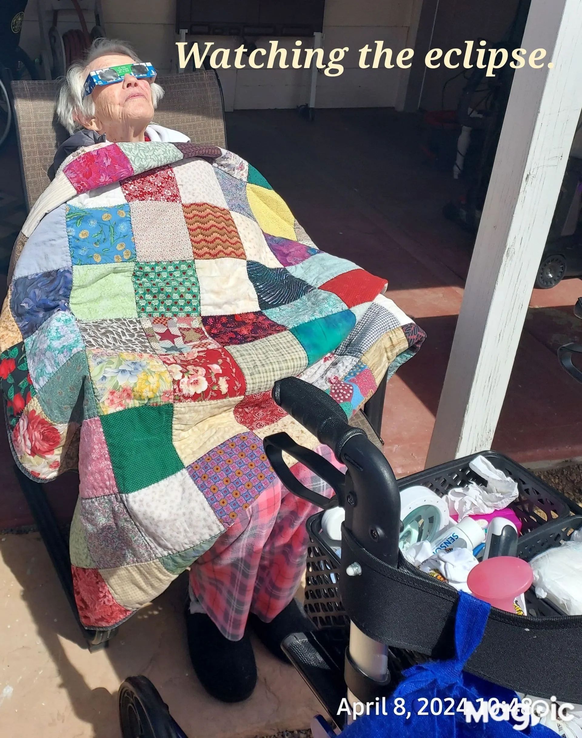 Elderly woman in sunglasses reclines in a chair, covered by a patchwork quilt, watching the eclipse on a sunny day.