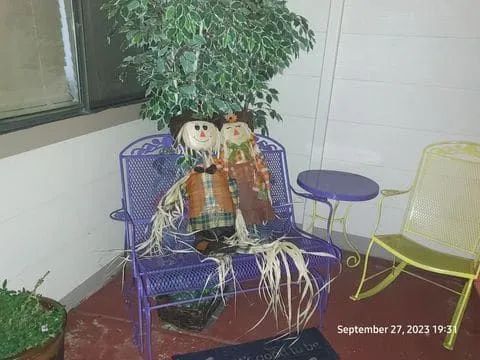Two scarecrows sit on a purple metal chair on a porch, with a small table and a yellow chair nearby.