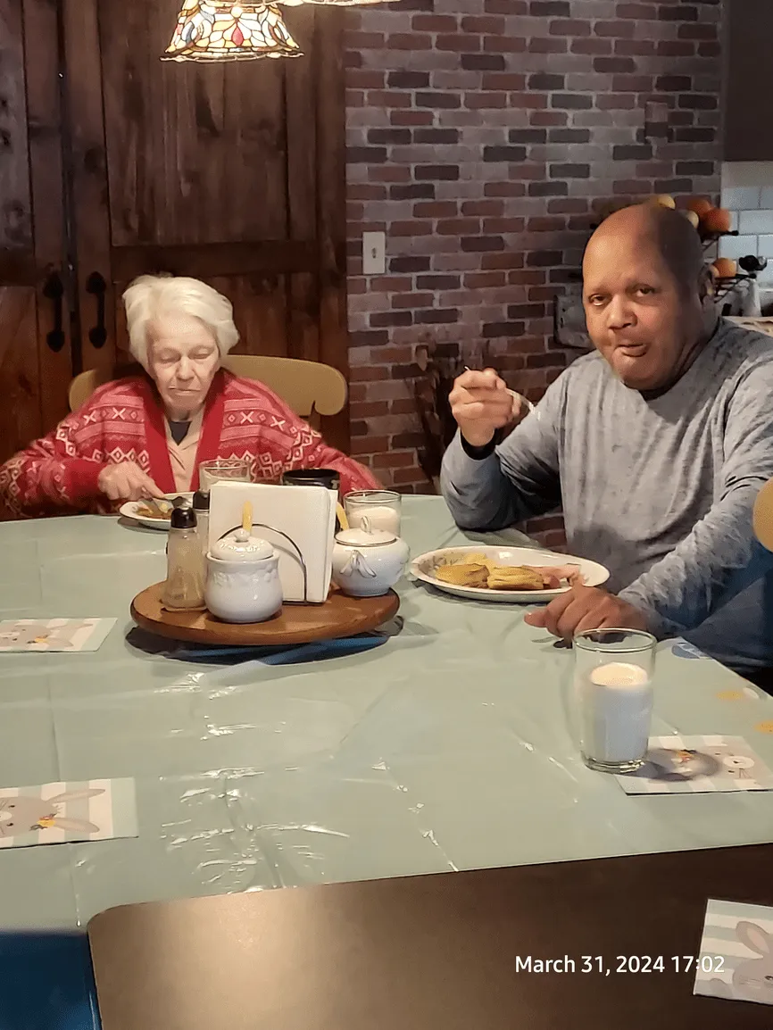 An older woman and a Black man eating at a table, in a dining room. The man holds a fork, smiling at the camera.
