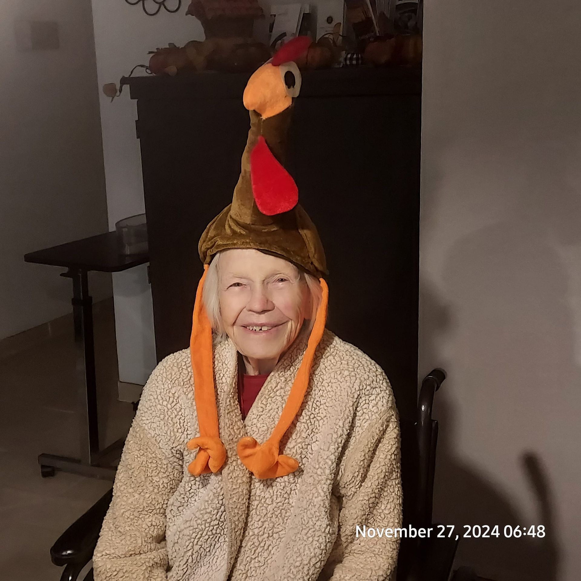 Elderly woman in a turkey hat smiles at the camera, wearing a fuzzy beige coat. The setting is indoors.