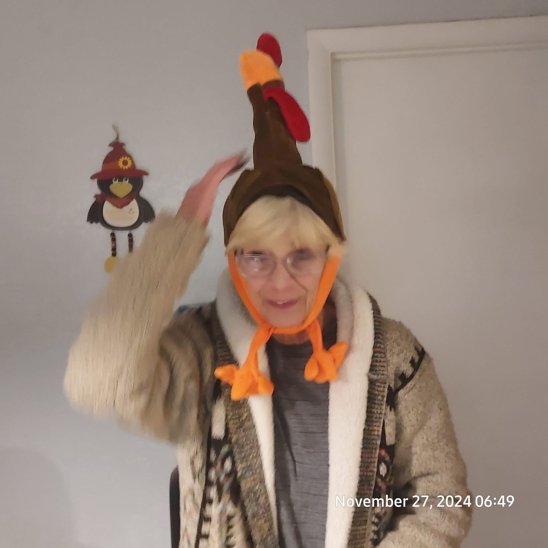 Person wearing a turkey hat, raising their hand, with a turkey decoration on the wall. The person is wearing a robe.