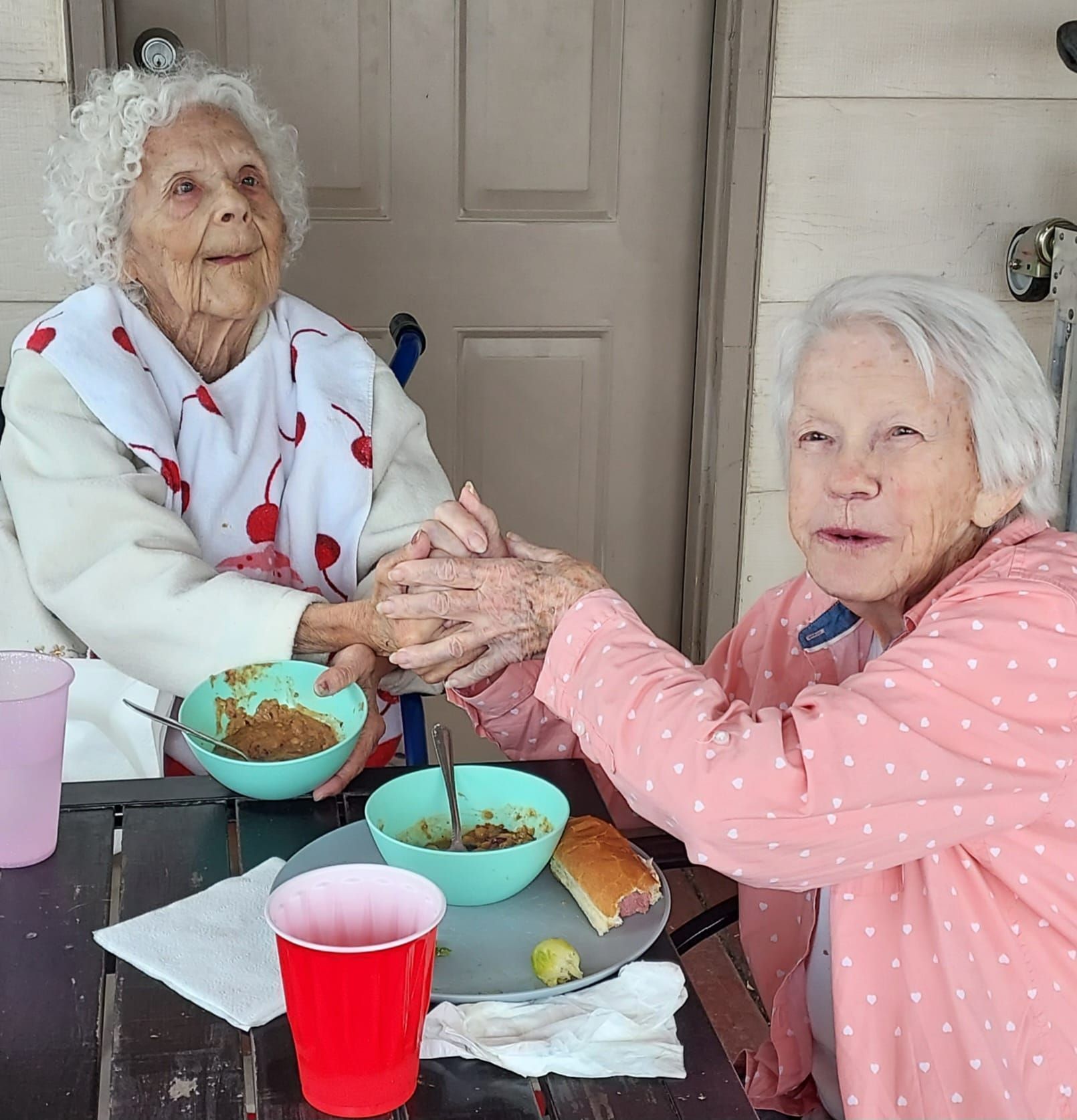 Two elderly women sitting outside, holding hands and eating soup. One wears a bib; both have smiles.