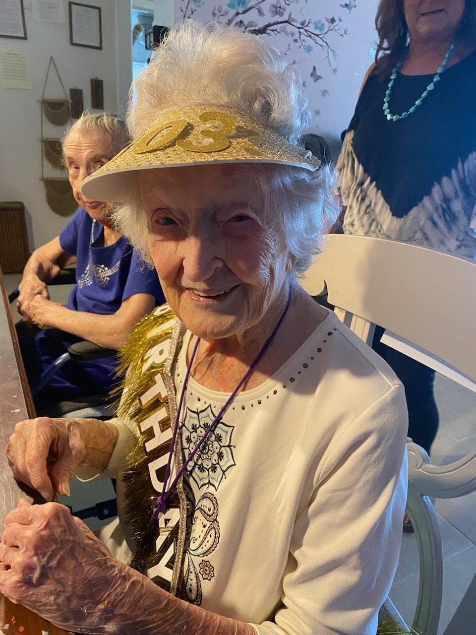 Elderly woman wearing a birthday hat and sash, smiling at the camera.