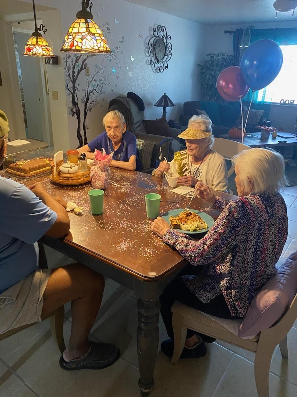 Four older women and another person eat at a table decorated with confetti; balloons are visible in the background.