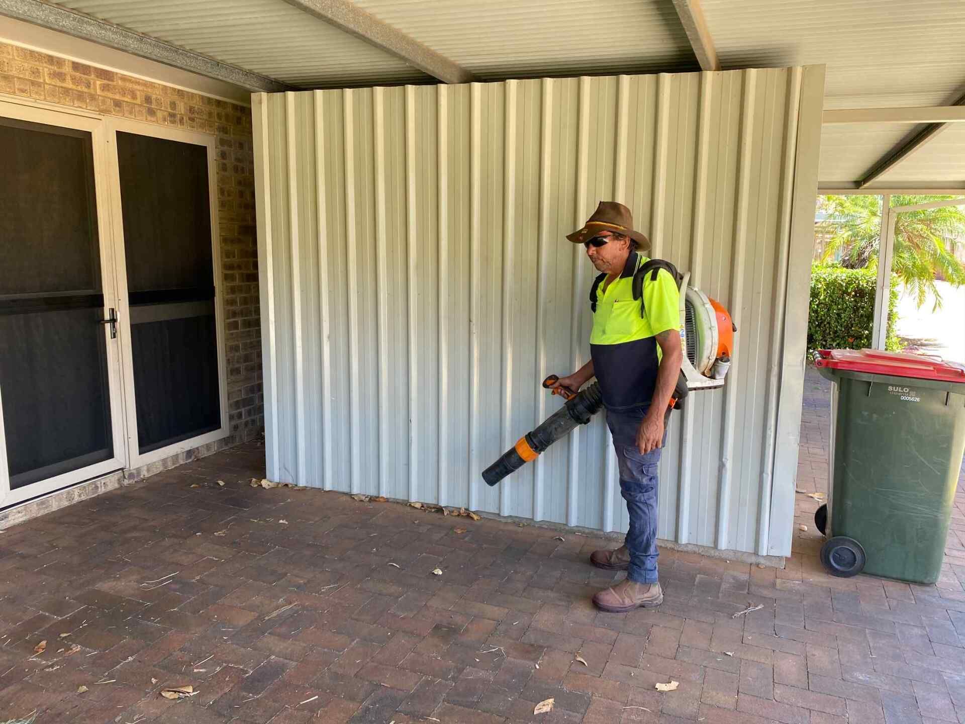 a Person Uses a Leaf Blower Near a Wall and Trash Can in a Covered Outdoor Area — Darrell's Mowing Services in Sunset, QLD