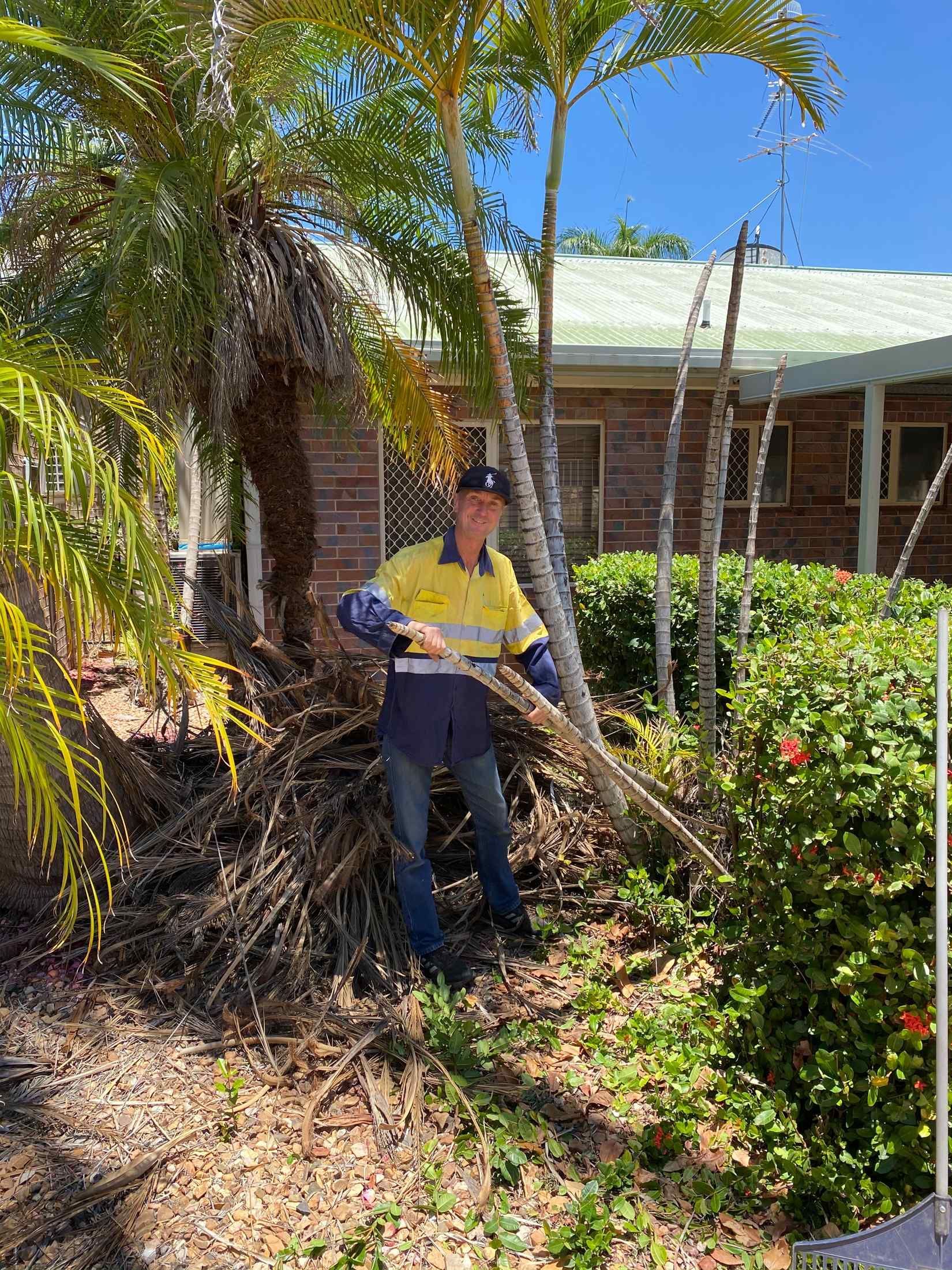Man Wearing Work Clothes Raking Leaves Near Palm Trees — Darrell's Mowing Services in Sunset, QLD