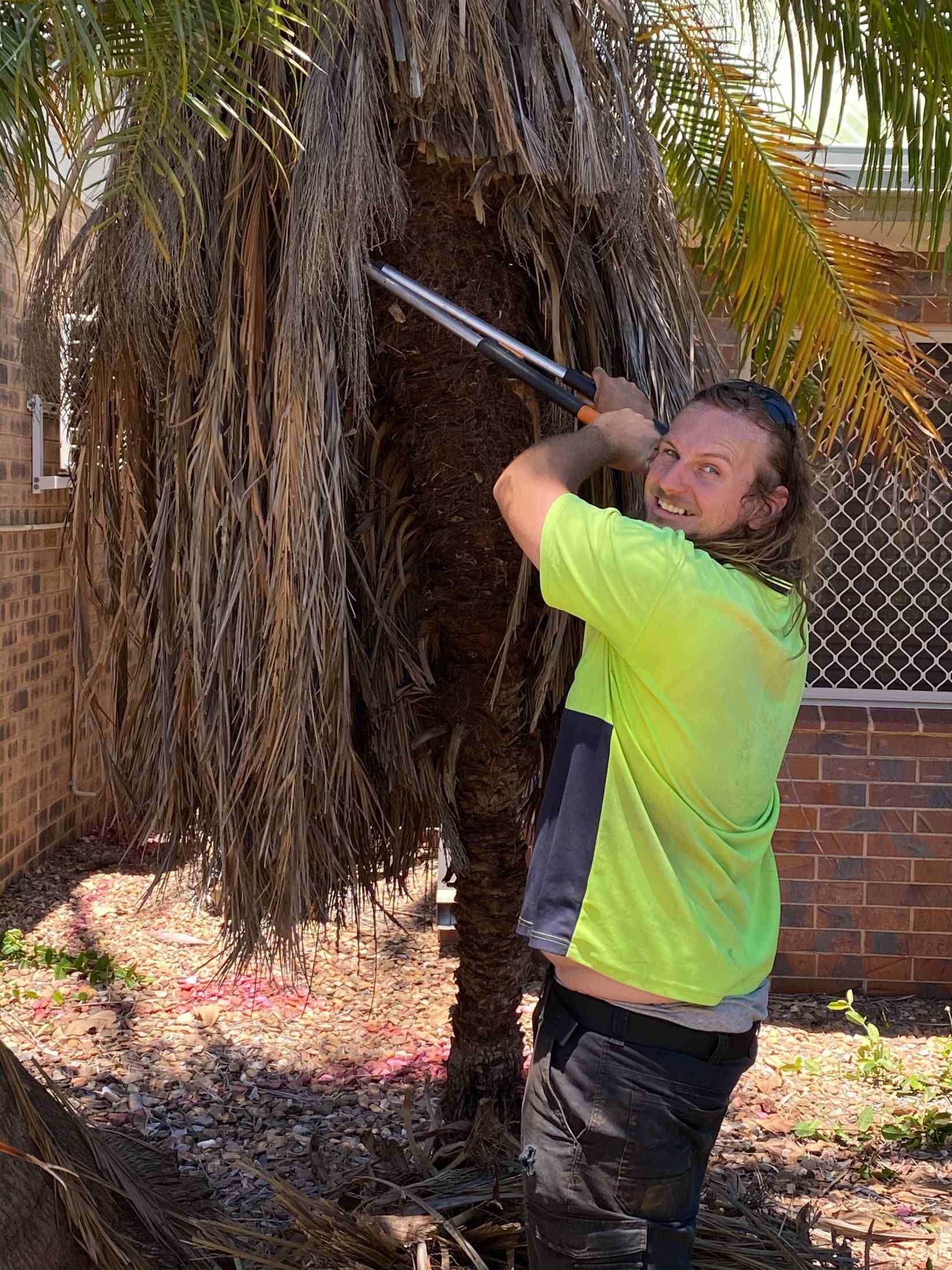 Man in Neon Shirt Sawing Palm Tree With a Metal Saw — Darrell's Mowing Services in Sunset, QLD