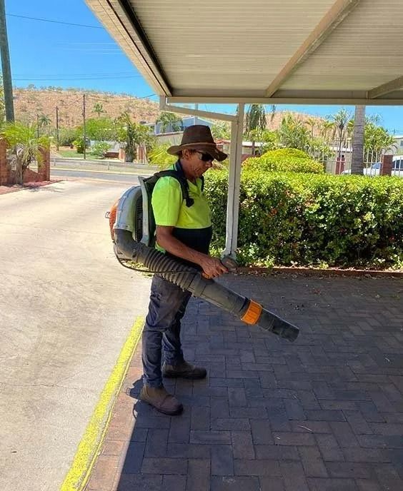 Man using a leaf blower on a paved area, wearing a hat and safety vest — Darrell's Mowing Services in Sunset, QLD