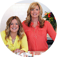 Two women are standing next to each other in a kitchen and smiling.