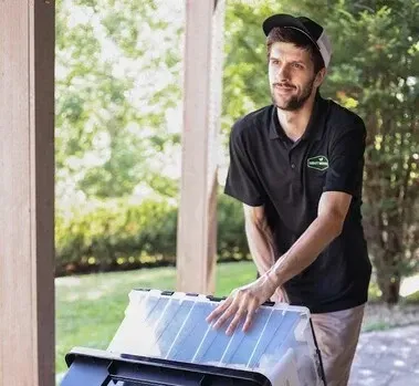 Man in black polo and cap pushing a rolling bin outside; greenery in background.