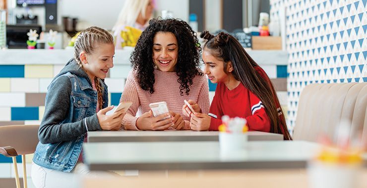 Three youth look at a phone together at a table in a cafe, smiling.