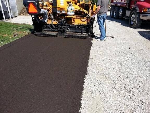 A paving machine lays fresh, dark asphalt on a light gravel driveway, with a worker standing nearby.
