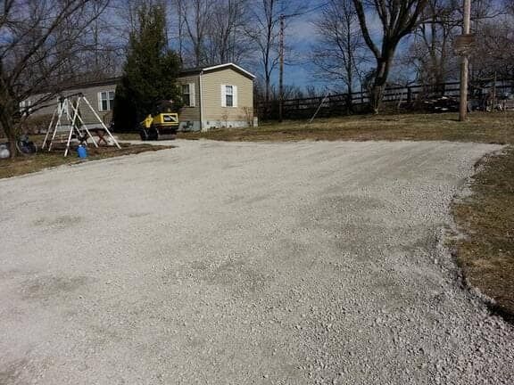 A gravel driveway leading to a mobile home on a sunny, clear day, with a swing set visible in the yard.