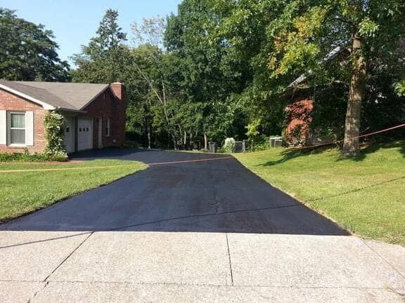 A freshly paved asphalt driveway leading to a brick house next to a grassy lawn and trees under a clear blue sky.
