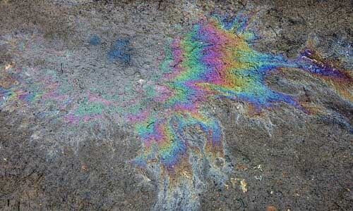 A rainbow-colored oil slick spreads across a patch of rough, gray concrete or wet ground. A rainbow-colored oil slick spreads across a patch of rough, gray concrete or wet ground.