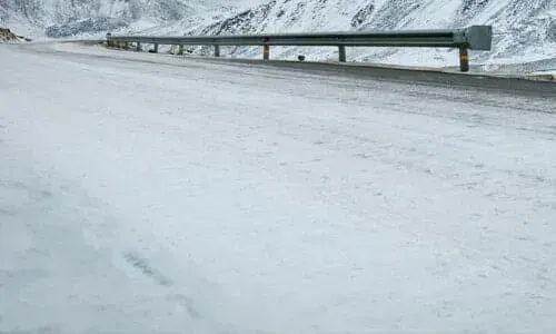 A snow-covered road bordered by a metal guardrail, with snowy mountains visible in the background. A snow-covered road bordered by a metal guardrail, with snowy mountains visible in the background.