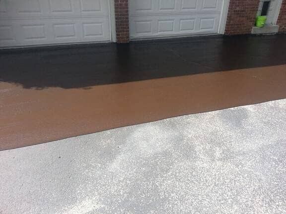 A partially seal-coated driveway with a distinct contrast between the dark brown fresh coating and the aged gray concrete.