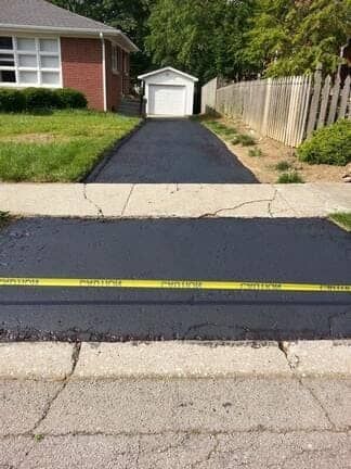 A freshly paved, black driveway blocked by a yellow caution tape in front of a residential house and garage.