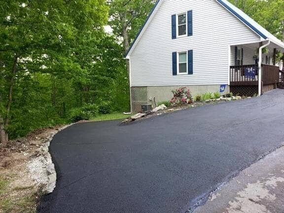 A freshly paved asphalt driveway leading to the side entrance and deck of a white house surrounded by trees.