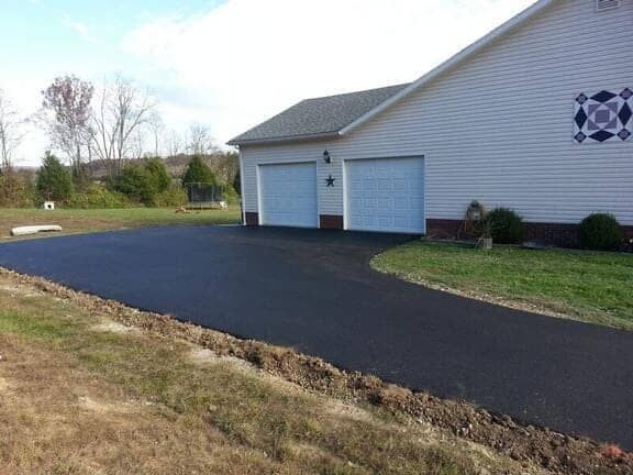 A newly paved black asphalt driveway leads to a white residential garage with two doors on a grassy lot.