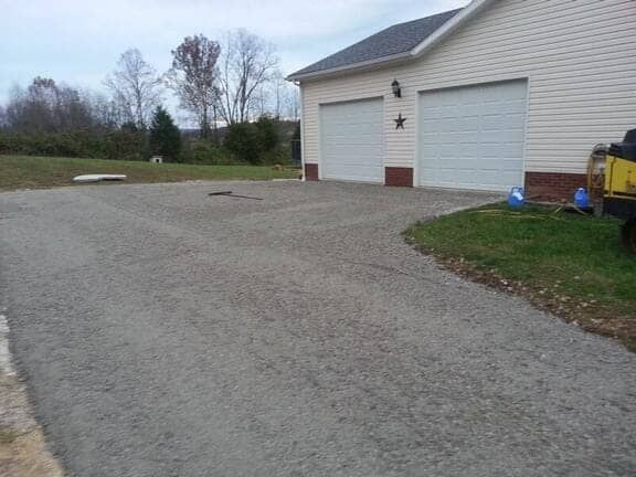 A light-colored house with a two-car garage sits beside a large gravel driveway surrounded by grass and trees.