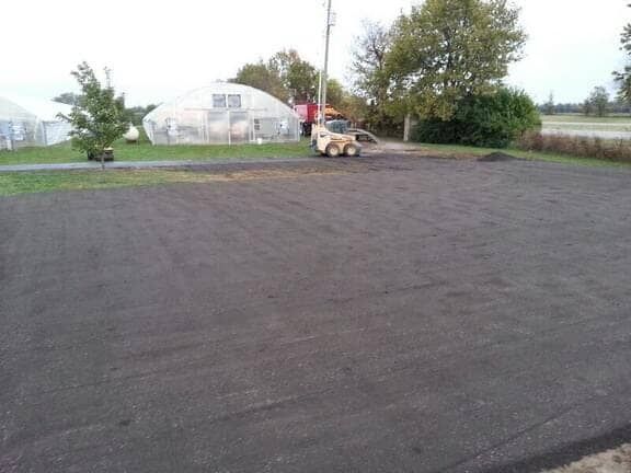 A construction vehicle parked on a large, freshly leveled patch of dark soil near a greenhouse and trees.