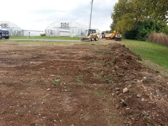 Construction equipment clears a dirt field in front of large greenhouses on an overcast day.