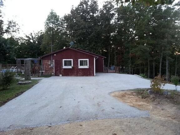 A red single-story house with a white-trimmed gravel driveway in front, surrounded by a forest.