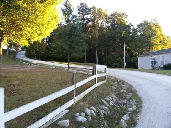 A gravel driveway curves past a white fence and a small house toward a tree-lined hill under a bright, sunny sky.