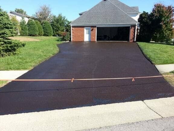 A newly sealed, dark asphalt driveway leads to a brick house with a two-car garage on a sunny day.
