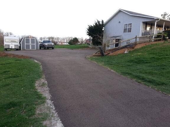 A paved driveway leads to a light blue house on the right, with a shed and parked vehicle in the background.